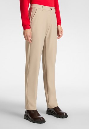 Pantalones ajustados en color beige con pliegues frontales, bolsillos laterales y cierre de botón, combinados con zapatos marrón oscuro. Textura de tela suave.