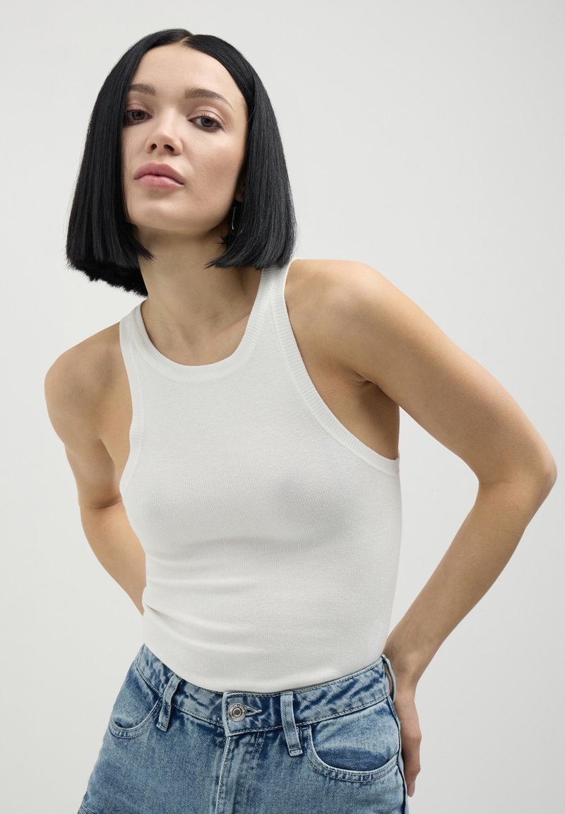 Débardeur blanc côtelé avec un col montant et de larges emmanchures, associé à un short en denim bleu taille haute avec une fermeture à bouton.