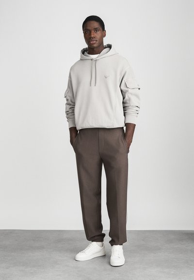 Bărbat îmbrăcat cu un hanorac bej deschis cu buzunar frontal, pantaloni jogger maro cu dungi laterale și adidași albi, stând cu mâinile în buzunare.