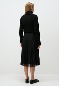Schwarzer Strickrollkragenpullover mit entspannter Passform, kombiniert mit einem schwarzen, plissierten Midikleid. Der Look wird mit schwarzen Ballerinas abgerundet.