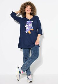 Marineblaues Tunika mit V-Ausschnitt und drei Viertelärmeln, verziert mit einem bunten Blumenmuster. Kombiniert mit blauen Jeans und mehrfarbigen Turnschuhen.