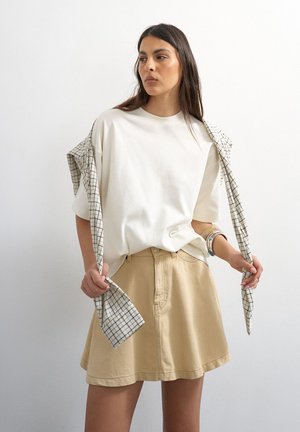 Tânără purtând un tricou alb lejer și o fustă bej, ținând o eșarfă în carouri drapată peste umeri, pe un fundal simplu.