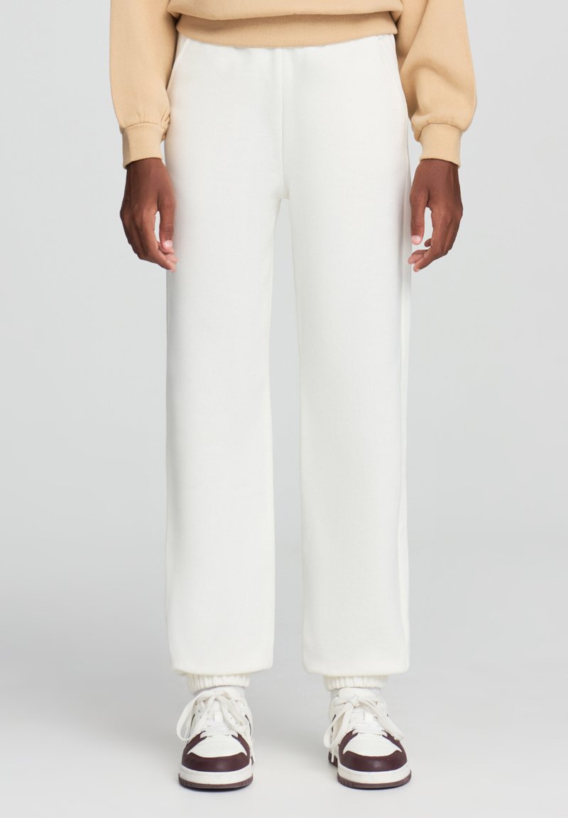 Białe dresowe spodnie o luźnym kroju, z elastycznymi mankietami i gładką fakturą, w połączeniu z białymi i brązowymi snekerami.