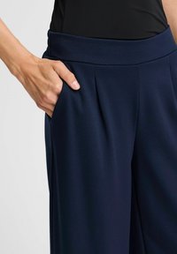 Tmavě modré kalhoty vyrobené z měkké tkaniny, s širokým pasem, sklady a bočními kapsami, které ukazují hladkou texturu a pečlivě propracovaný design.