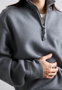 Pull gris à fermeture éclair au quart, fabriqué en tissu doux. Il présente un col haut, des poignets côtelés et une coupe décontractée, mettant en valeur une texture lisse.