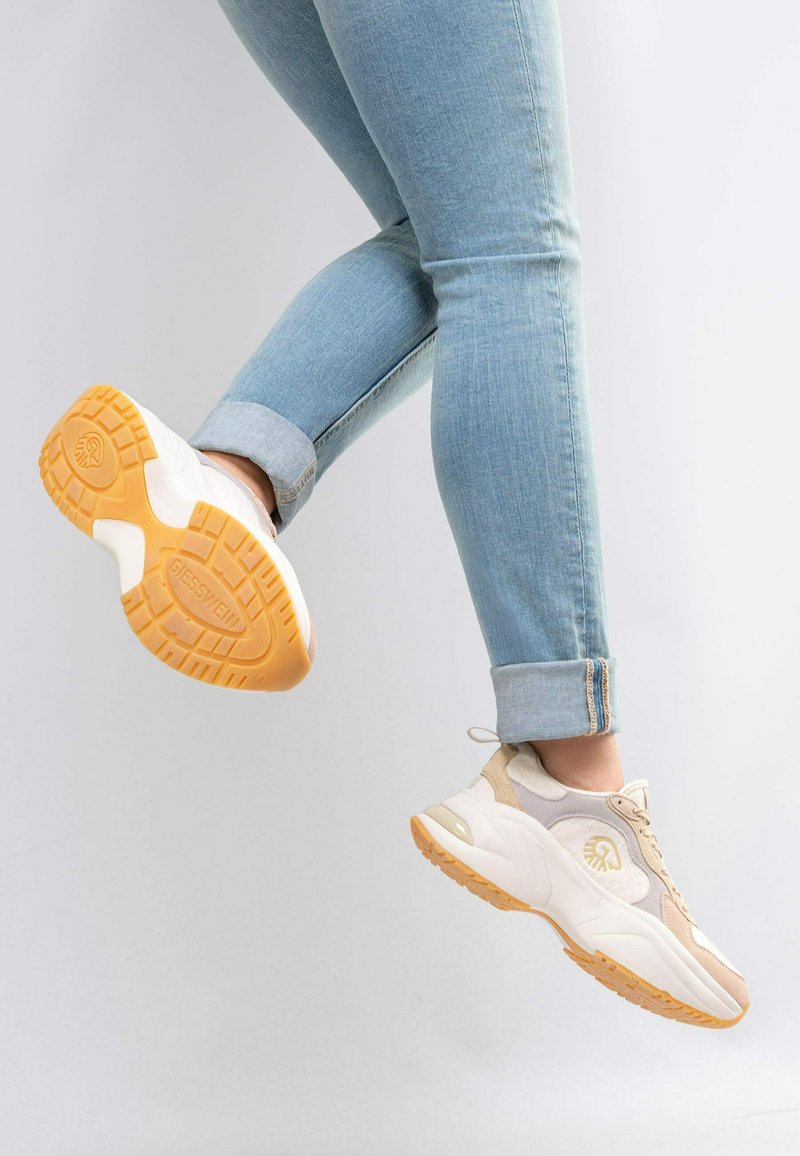 Benen in opgerolde lichtblauwe spijkerbroeken met witte en beige sneakers met oranje zolen, één voet midden in de lucht geheven tegen een effen achtergrond.