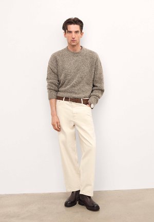 Maglione beige lavorato a maglia, pantaloni crema, cintura in pelle marrone e stivaletti alla caviglia marrone scuro. Modella in piedi con le mani in tasca su uno sfondo neutro.