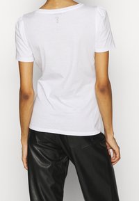 Camiseta de algodón blanca con mangas cortas, costuras de hombros ligeramente abullonadas y un pequeño detalle gráfico en la parte trasera. Combinada con pantalones de cuero negros.