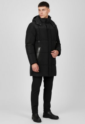 Mann trägt eine schwarze, gesteppte Winterjacke mit Kapuze und Reißverschlusstaschen, schwarze Hose und schwarze Stiefel, steht vor einem schlichten grauen Hintergrund.