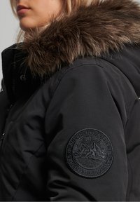 Černá parka s kapucí lemovanou kožešinou, prošívanou texturou a kulatou černou nášivkou na levém rukávu s raženou značkou.