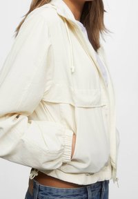 Veste légère crème avec capuche à cordon de serrage, poignets élastiques, poches latérales et coupe décontractée. Le tissu texturé apporte un détail subtil.