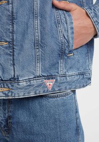Denim jacket in light blue, featuring a triangle logo patch in red and white, distressed details, and classic jean jacket cut with pockets.