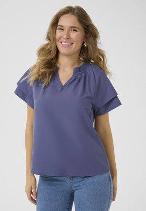 Femme souriante aux longs cheveux ondulés portant des boucles d'oreilles argentées en forme d'anneau, un chemisier violet à manches courtes et un jean bleu, posant devant un fond uni.