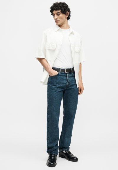 Άνδρας που φοράει λευκό κοντομάνικο πουκάμισο πάνω από λευκό μπλουζάκι, μπλε τζιν με μαύρη ζώνη και μαύρα παπούτσια, στέκεται με το ένα χέρι στην τσέπη.