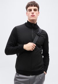 Νέος άνδρας με κοντά καστανά μαλλιά, που φοράει μαύρο μπουφάν με φερμουάρ και γκρι παντελόνι, κρατώντας έναν μαύρο ιμάντα τσάντας πάνω στον ώμο του.