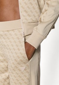 Μπεζ φόρμα με υφή και επαναλαμβανόμενο μοτίβο λογότυπου. Ζακέτα με φερμουάρ, πλαϊνή τσέπη και ριμπ μανσέτες, με λεία υφασμάτινα στοιχεία.