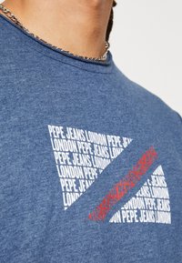 Camiseta azul con el texto repetido "PEPE JEANS LONDON" en blanco formando un triángulo, cruzada por una franja roja con un texto similar superpuesto.