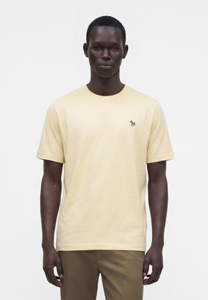 Homme portant un t-shirt jaune clair à manches courtes avec un petit logo zèbre coloré sur la poitrine gauche et un pantalon marron.