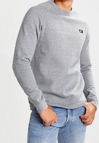 Harmaa huppari, jossa on pyöreä kaula-aukko, raglan-hihat ja pieni musta logopatchi rinnassa, yhdistettynä sinisiin farkkuihin.