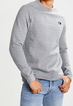 Felpa grigia con colletto rotondo, maniche raglan e una piccola patch nera sul petto, abbinata a jeans blu.