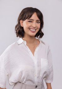 Weißes, leicht zerknittertes Button-up-Hemd mit einer Brusttasche auf der linken Seite. Kurze Ärmel, lockere Passform. Trägt kleine goldene Schmetterlingsohrenringe.