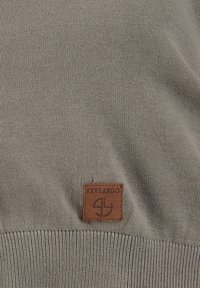 Lichtbruin gebreide stof met een klein rechthoekig bruin patch met het label "KEY LARGO" en een ronde logo onder de tekst.