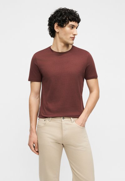Homme aux cheveux bouclés portant un t-shirt bordeaux à manches courtes et un pantalon beige, debout avec une main dans la poche, regardant vers la droite.