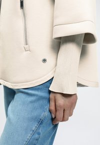 Ανοιχτό μπεζ φούτερ με φερμουάρ μπροστά και φαρδιά μανίκια, συνδυασμένο με ανοιχτό μπλε τζιν. Διαθέτει ριμπ μανσέτα και κυκλικό λογότυπο της μάρκας.