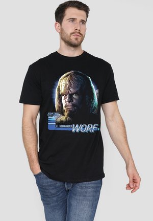 Man wearing black Star Trek T-shirt featuring Lt. Commander Worf's face and name, paired with blue jeans, standing against gray background.