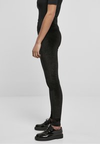Person som bär svarta sammetshöga leggings, svart kortärmad topp och blanka svarta snörskor, står i profil mot en enfärgad bakgrund.
