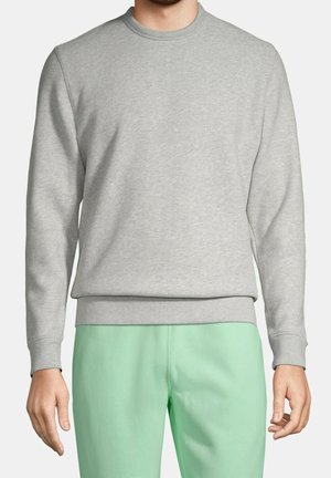 Homme portant un sweat-shirt gris clair à col rond et un pantalon vert clair, debout, bras détendus le long du corps, sur un fond neutre.