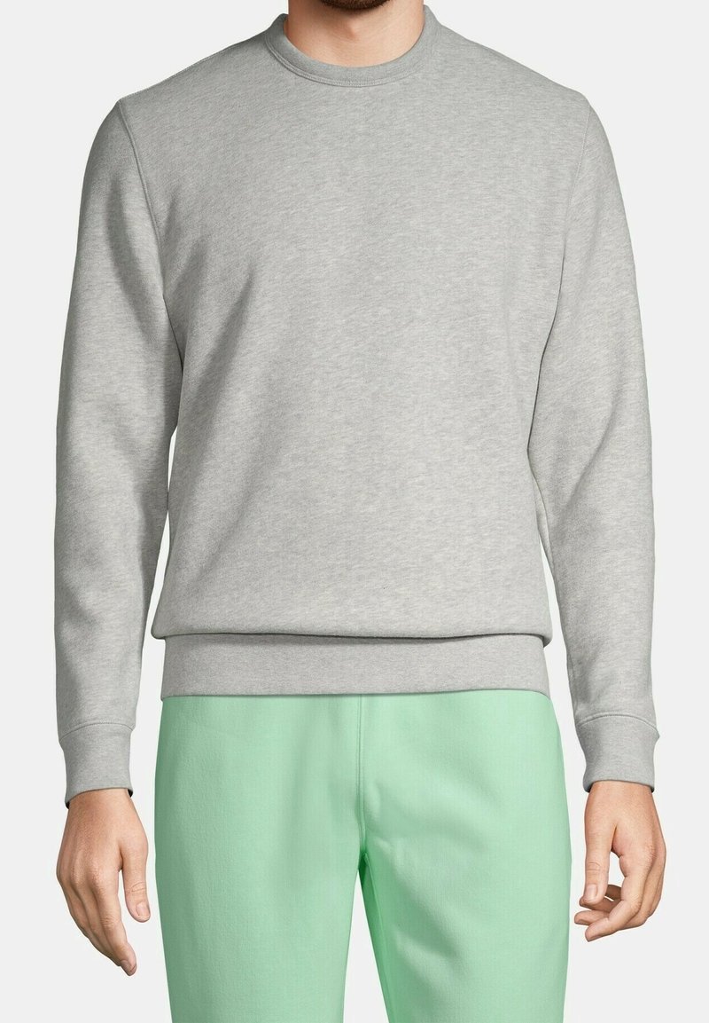 Homme portant un sweat-shirt gris clair à col rond et un pantalon vert clair, debout, bras détendus le long du corps, sur un fond neutre.