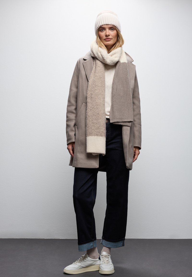 Abrigo de lana beige sobre un suéter color crema, combinado con una bufanda a juego. Jeans de mezclilla oscuros y zapatillas blancas. Un gorro de punto texturizado completa el atuendo.
