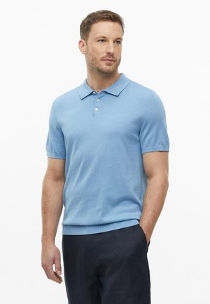 Homme portant un polo bleu clair à manches courtes et un pantalon foncé, debout avec une main dans la poche, regardant sur le côté sur fond blanc.