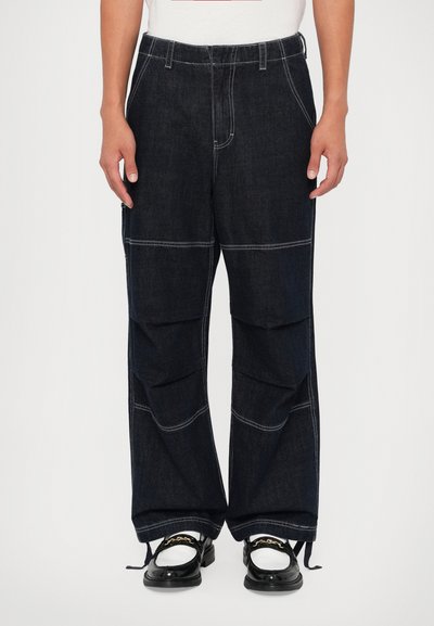 Pantalons en denim bleu foncé avec une coupe ample, couture visible, poches latérales et ourlets à cordon de serrage réglables. Portés avec des chaussures noires.