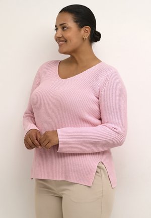 Lys pink strikket sweater med V-hals, lange ærmer, sideslidser og et mønstret design, parret med brune bukser.