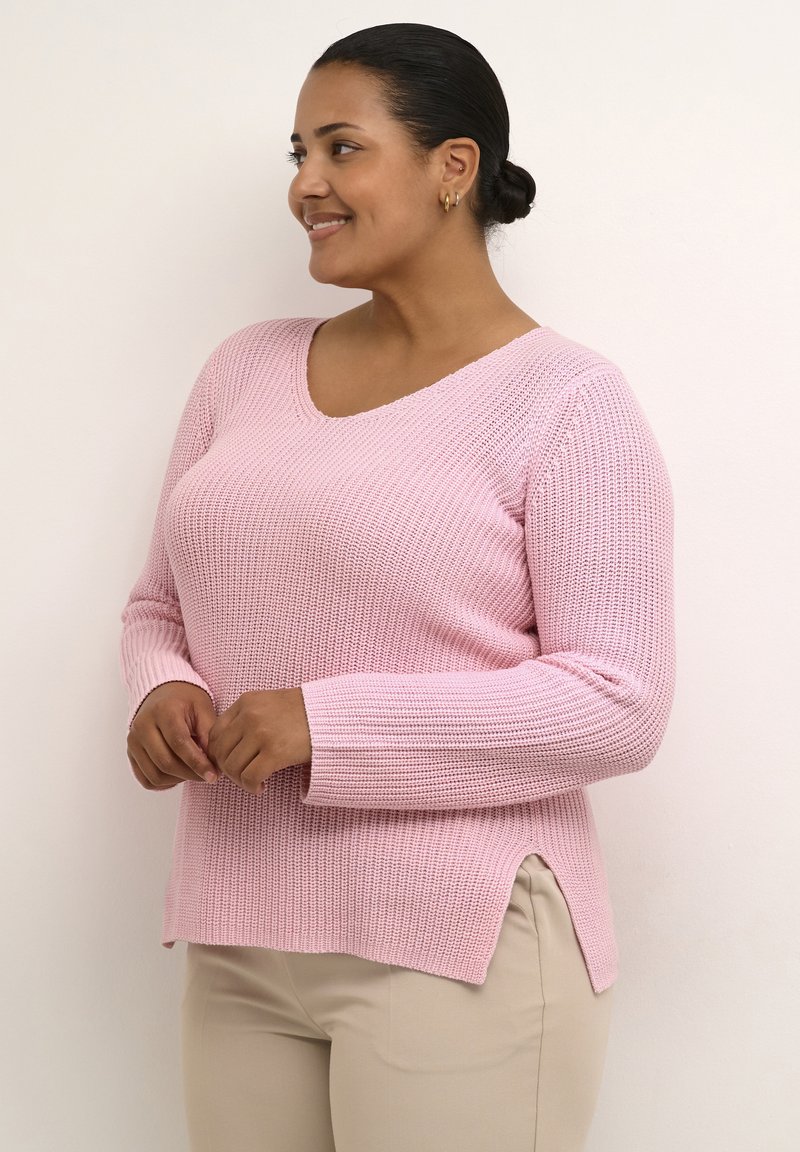 Pull en maille rose clair avec un col en V, des manches longues, des fentes latérales et un motif texturé, assorti à un pantalon beige.