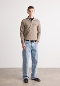 Camisa polo de manga longa castanha com colarinho em contraste, combinada com jeans de perna larga azul claro e sapatos pretos. Modelo está de pé contra um fundo neutro.
