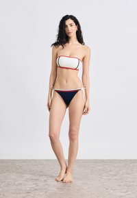 Σετ μπικίνι με κρεμ τοπ bandeau με κόκκινα και μαύρα στοιχεία, και ναυτικό κάτω μέρος χαμηλής μέσης με κορδόνια στα πλαϊνά και κορδόνια στο ίδιο χρώμα.