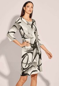 Schwarz-weißes Kleid mit abstraktem Muster, dreiviertellangen Ärmeln, figurnaher Taille und einer Knoten-Detail am Bund, gefertigt aus einem glatten, elastischen Stoff.