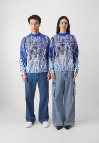 Blå og hvite mønstrede sweatshirt med elastiske mansjetter, kombinert med løs passform på denimbukser. Designet har intrikate svlinger og tekst.
