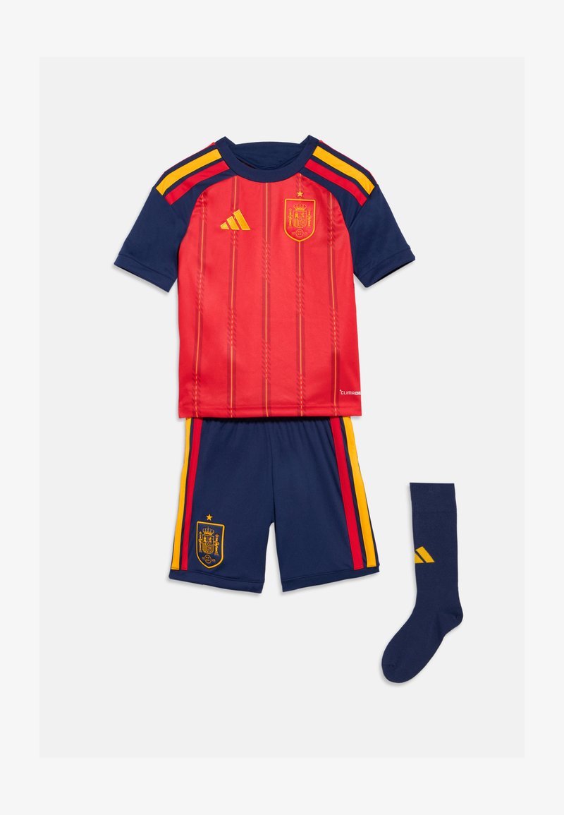 Camisola de futebol vermelha e azul-marinho com riscas amarelas e vermelhas, calções azuis-marinho combinando com riscas, e uma meia azul-marinho com um logótipo amarelo.