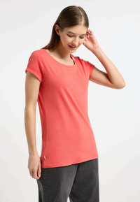 T-shirt corallo a maniche corte realizzato in tessuto morbido, con scollatura rotonda, dettagli decorativi con bottoni sulla spalla e spacchi laterali. Abbinato a pantaloni grigio scuro.