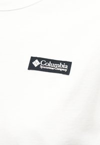 Μπατς με το λογότυπο της Columbia Sportswear Company σε λευκό κείμενο πάνω σε μαύρο ύφασμα, ραμμένο σε απλό λευκό ύφασμα.