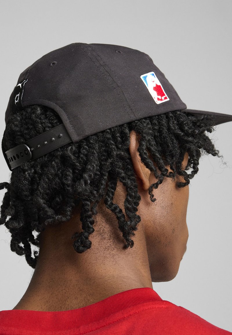 Cască de baseball neagră cu logo-ul NBA brodat, cu închidere ajustabilă tip snapback și vârf curbat, purtată într-un unghi, cu părul texturat vizibil.