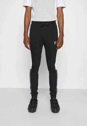 Man wearing zwarte joggers met witte en turquoise zijstrepen, zwarte sportschoenen, en een wit-grijs T-shirt met korte mouwen, staand tegen een witte muur.