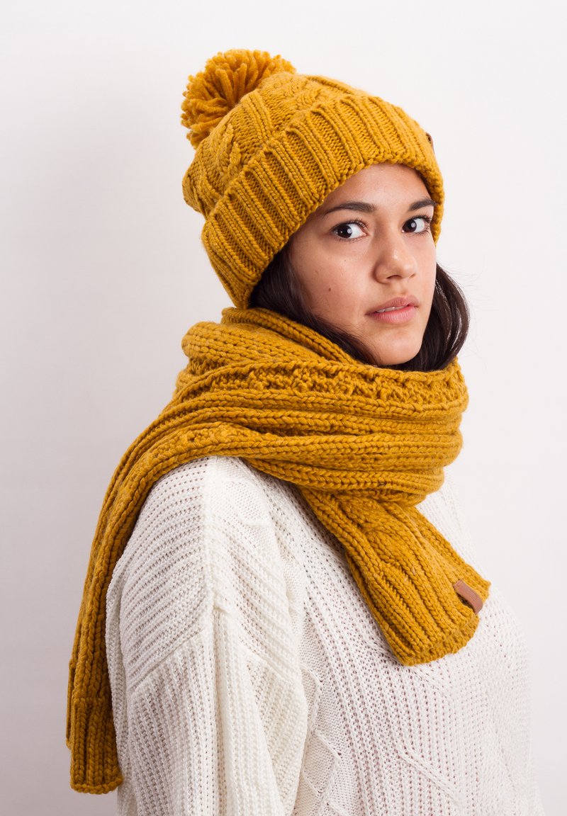 Knitted mustard yellow beanie with pom-pom and matching scarf, featuring a textured pattern and a leather accent. Worn over a white sweater.