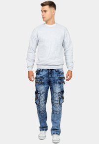 Felpa grigia con polsini a costine, abbinata a jeans cargo blu sbiaditi dotati di numerose tasche con zip e cuciture a rombo. Sneakers bianche.