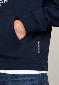 Sudadera azul marino con un bolsillo delantero, puños y dobladillo acanalados. Presenta una pequeña etiqueta de logo con acentos en rojo, blanco y azul en el lateral.