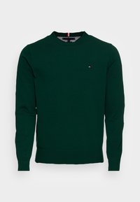 Pull en coton vert foncé avec des manches longues, un col rond et un petit patch logo rouge et blanc sur la poitrine gauche. Poignets et ourlet côtelés.